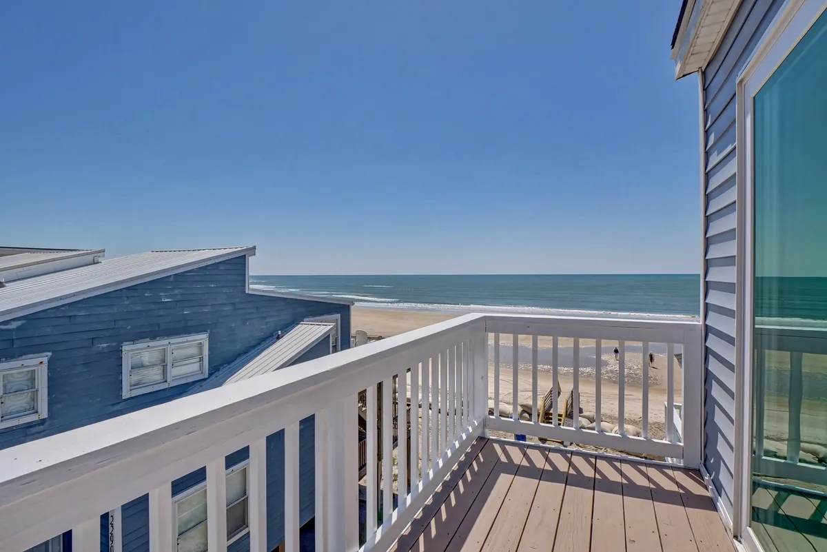 An exterior view from a balcony, showcasing a serene ocean vista framed by a simple railing and neighboring beach houses. The setup suggests a peaceful spot for morning coffee or stargazing at night.