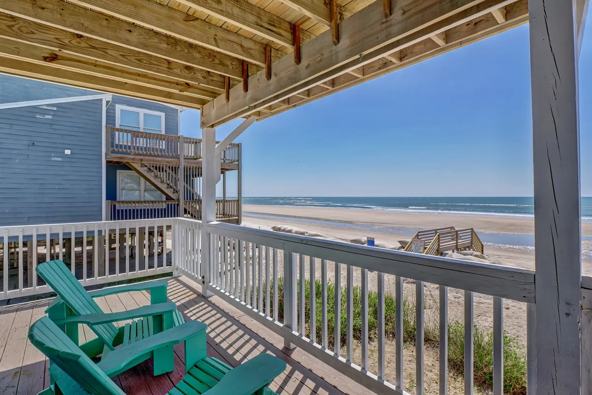 This photo captures a tranquil wooden deck furnished with green chairs overlooking a sandy beach and the ocean. The deck is spacious, open to the sea breeze, and safely enclosed with a railing, with easy access to the shore visible in the background.