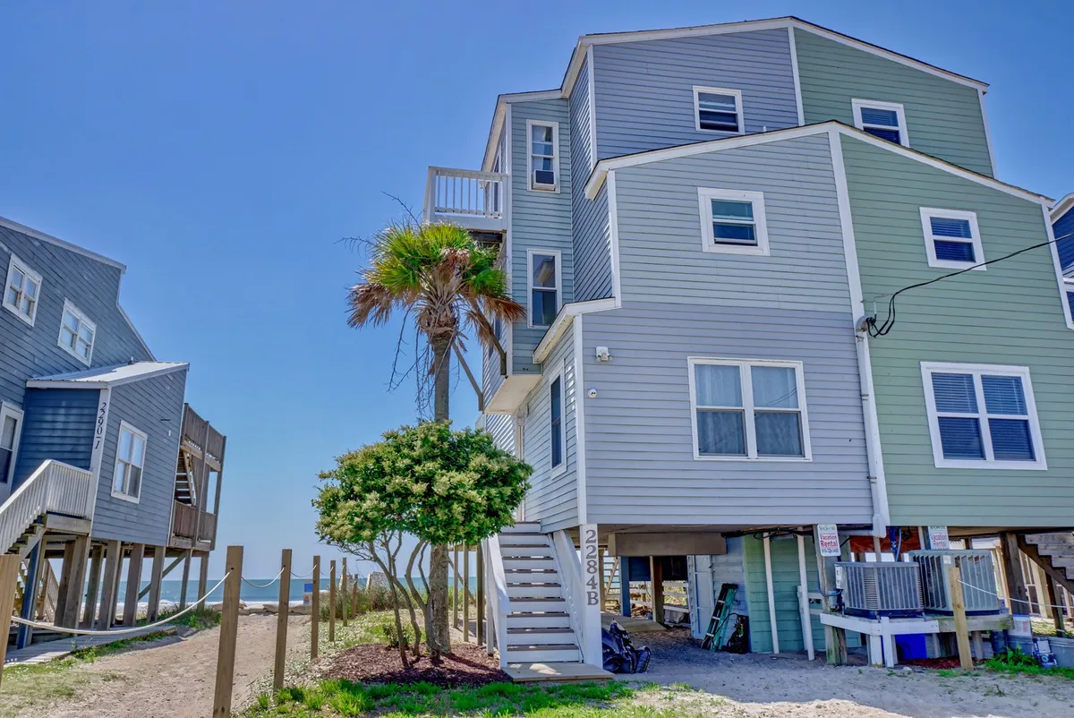 A multi-story beach house stands just steps from the shore, with a palm tree and a staircase leading to the entrance. The scene is bathed in soft sunlight, suggesting opportunities for sunset dinners on the balcony and memorable family gatherings.