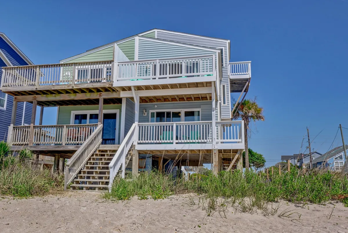 A cheerful wooden deck adorned with vibrant blue and green accents overlooks the sandy foreground leading to the beach. The open and spacious design invites guests and their pets to relax, socialize, and watch the waves together.
