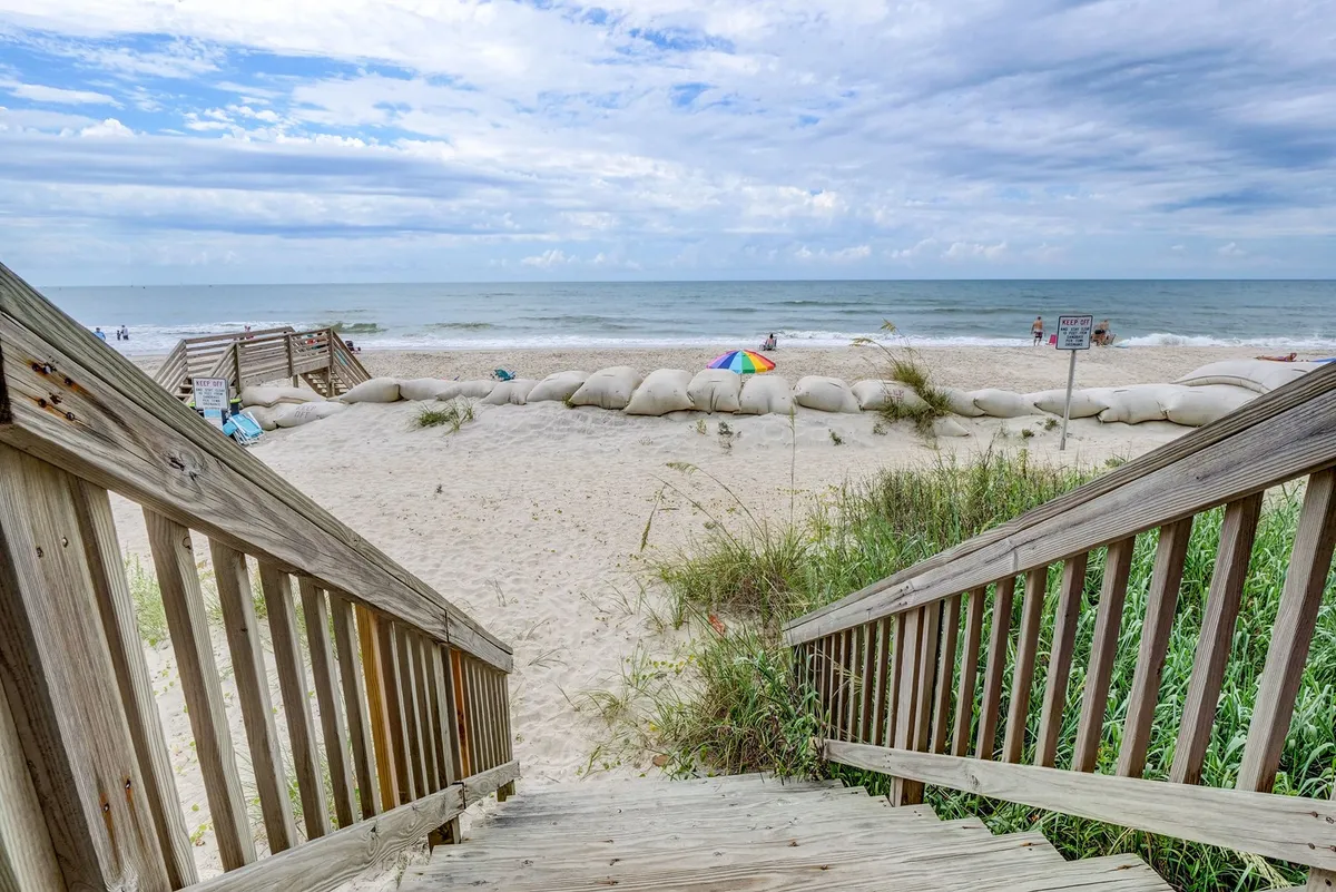 This image features wooden stairs leading directly down to the sandy beach, with the ocean visible beyond and a colorful umbrella set up on the sand. Sandbags line the path, and the wide expanse of beach is perfect for family activities like building sandcastles and beachcombing.