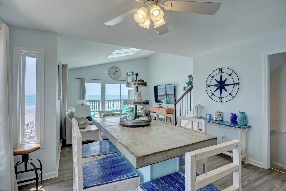 A large dining table sits by expansive windows overlooking the ocean. Nautical accents and a blue-and-white color palette create a casual coastal vibe, while the scene feels inviting for family meals or social gatherings.