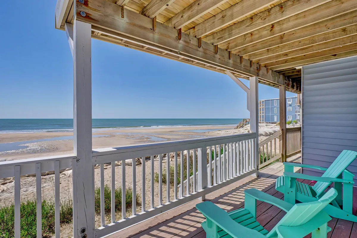 The image captures a serene oceanfront scene with a spacious wooden deck overlooking the Atlantic. Adirondack chairs invite guests to relax and unwind while taking in sweeping views of the beach and sky. The tranquil mood suggests a perfect setting for sunrise coffee, sunset cocktails, or stargazing.