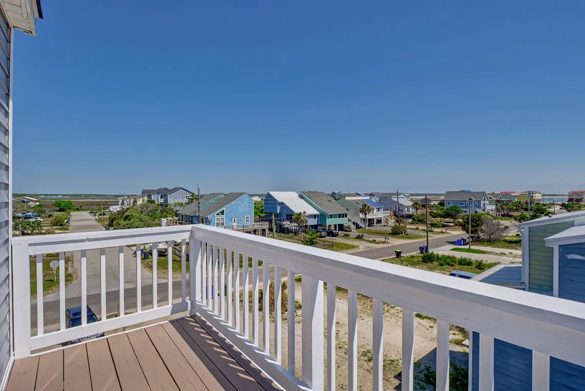 A tranquil view from a balcony, featuring a railing overlooking surrounding homes, a road leading toward the beach, and the distant shimmer of the ocean. The soft, early morning light enhances the sense of peaceful anticipation.