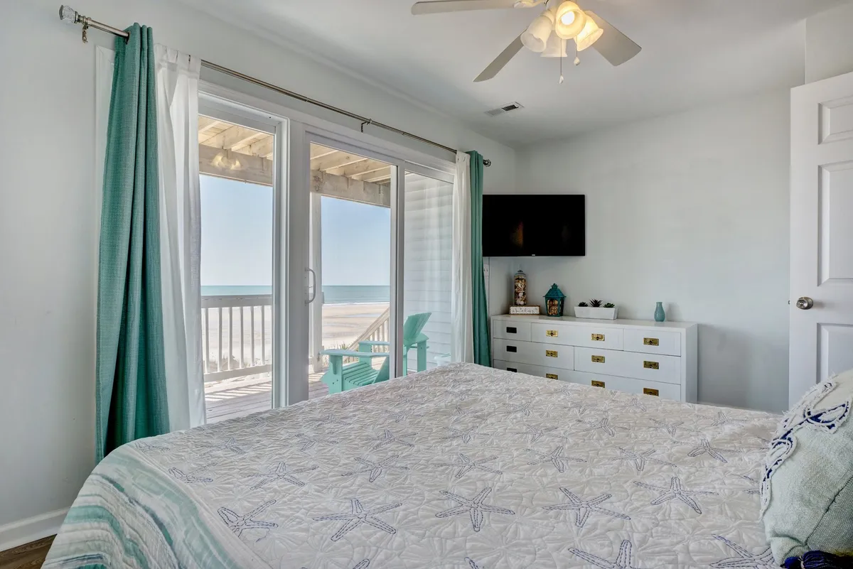 A serene master bedroom featuring a king bed with starfish-patterned bedding, large window with an ocean view, sliding glass doors that open onto a private deck, and elegant white furniture accented with gold. The setup is inviting, providing both a tranquil sleeping space and direct access to a secluded outdoor retreat.