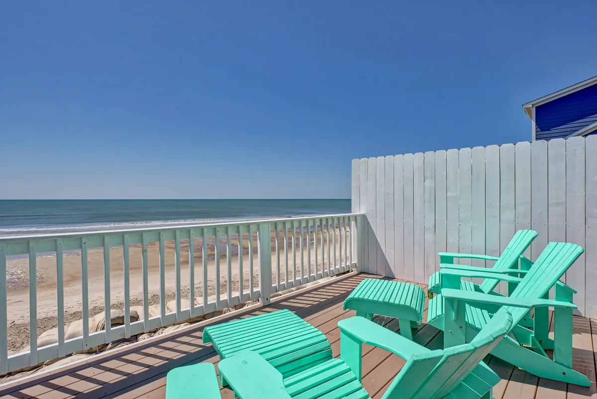 Turquoise Adirondack chairs are arranged on a deck overlooking the ocean, with a white railing framing the breathtaking view of the sand and surf. The relaxed seating invites families to enjoy sunrise coffees or unwind after a day on the beach.