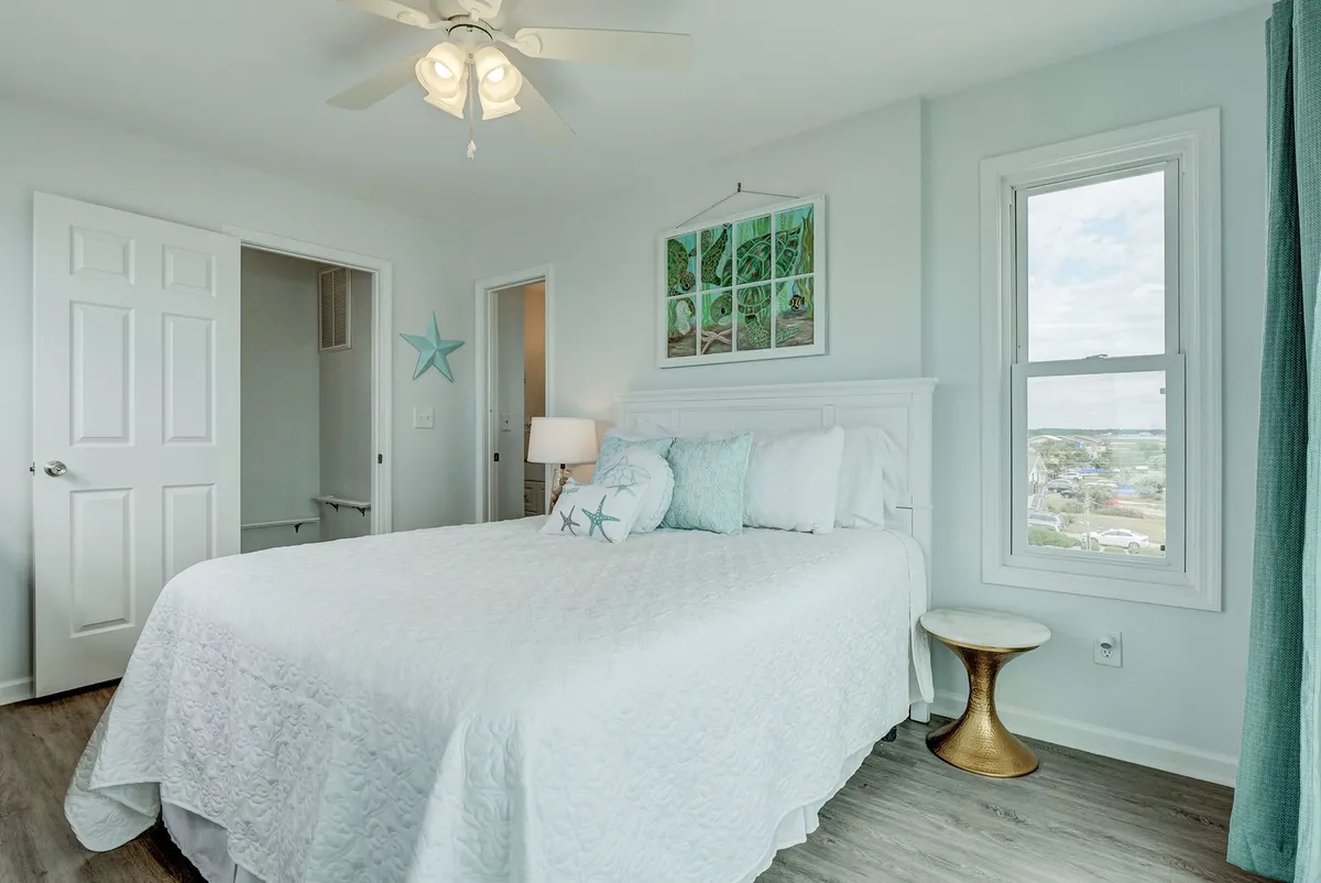 A cozy bedroom with a white quilted bed, decorative pillows, subtle starfish decor, and a window that lets in gentle natural light. The setting is calm, with inviting textures and a peaceful palette, suggesting a restful atmosphere ideal for relaxation and sleep.