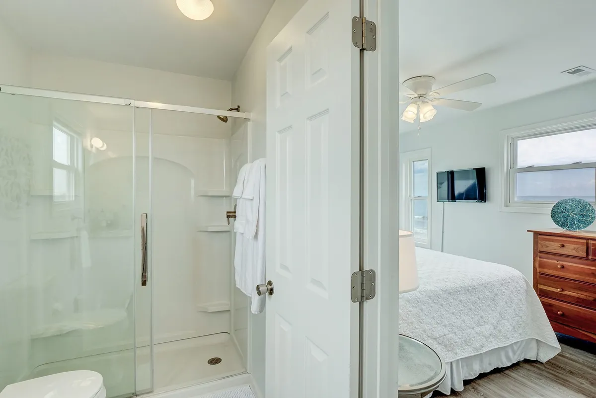 A serene bathroom scene with a glass shower enclosure, white towels thoughtfully hung, and a glimpse of the adjacent bedroom. The space is clean, well-organized, and offers a tranquil place to refresh and care for linens and towels.