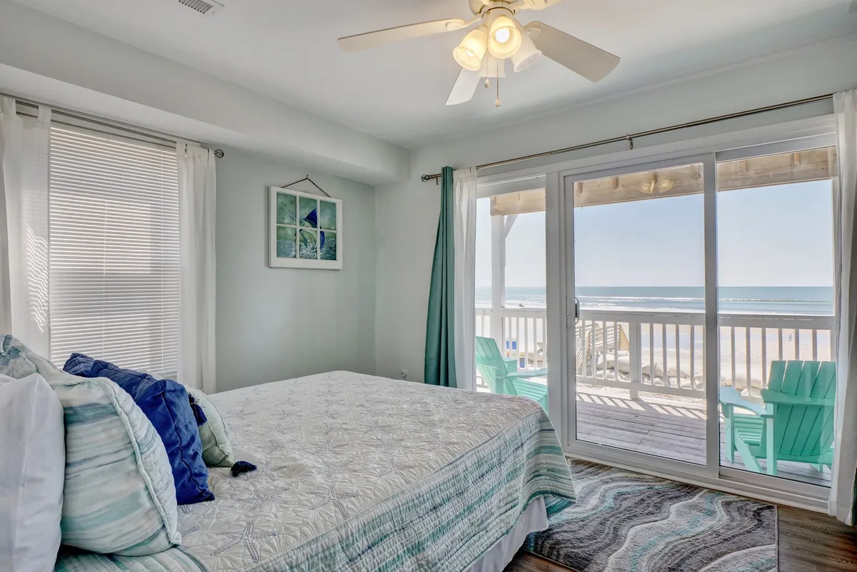 An inviting bedroom featuring a bed with decorative pillows and a large glass sliding door that opens directly onto a deck with beach chairs. The view reveals the ocean beyond, connecting the interior comfort with the outdoor, pet-friendly space.