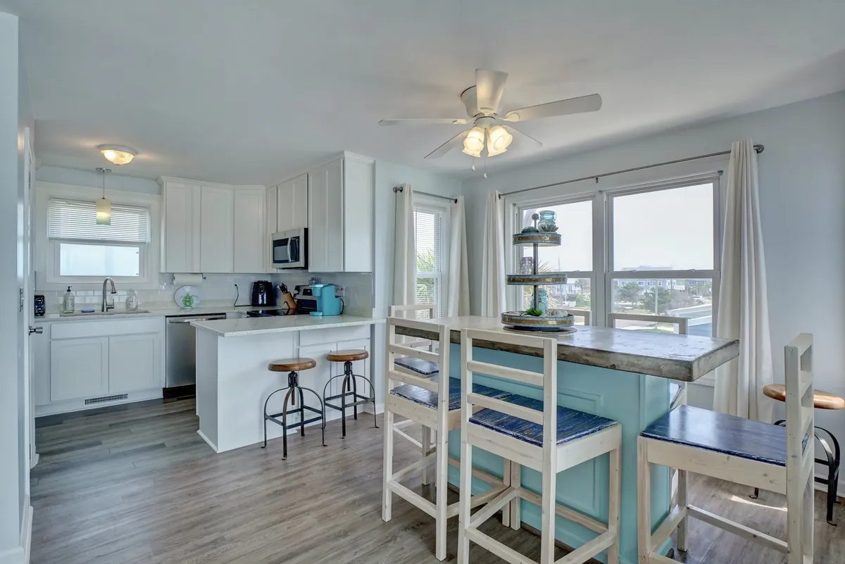 The kitchen area is bright and welcoming, with a spacious island, comfortable bar stools, and windows that let in streams of sunlight. The layout encourages socializing and casual dining.