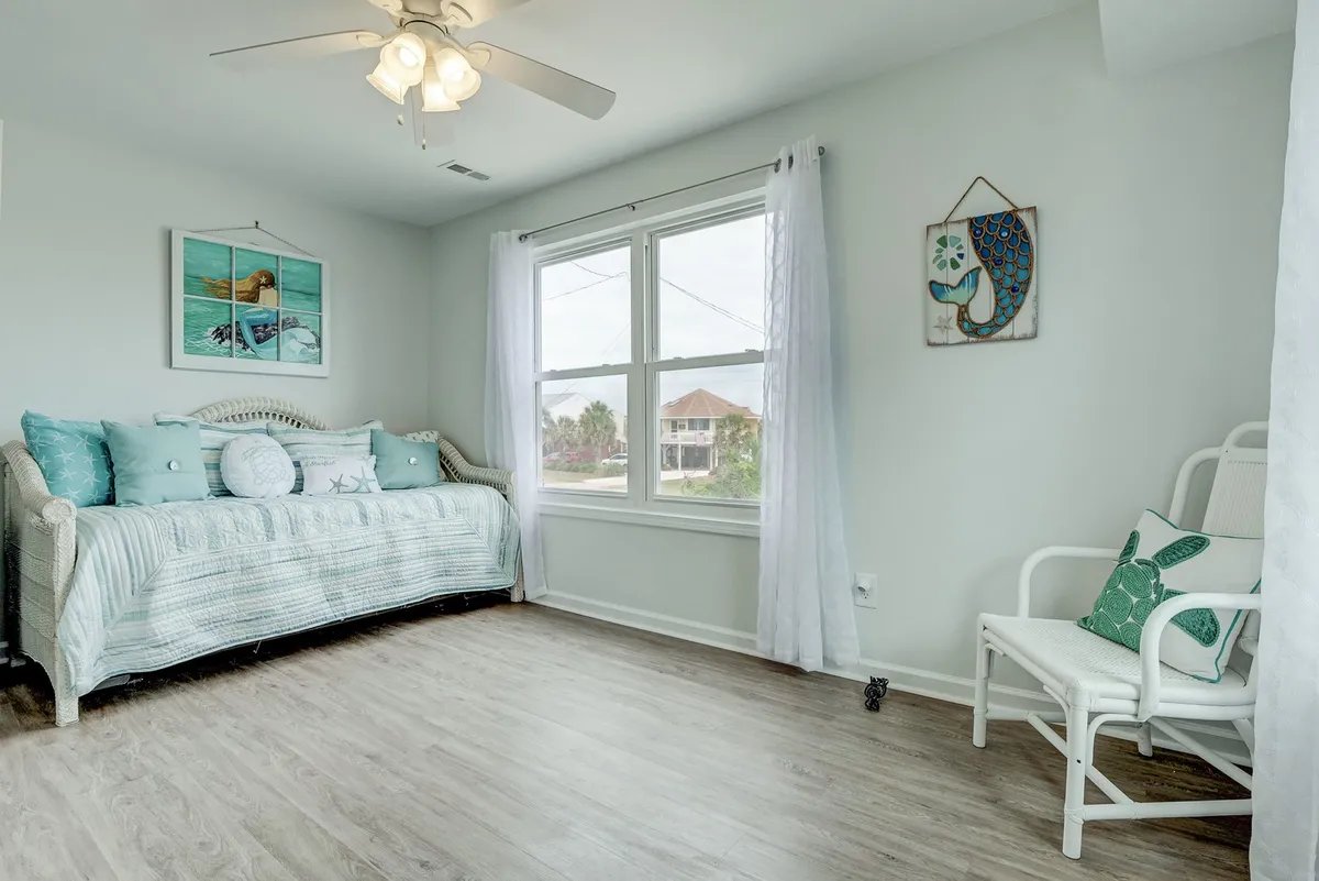 A cheerful twin room featuring a daybed with decorative pillows, a large window draped with sheer curtains, and a small accent chair. The space is bright and welcoming, ideal for families or friends, and offers a flexible sleeping arrangement with a trundle under the daybed.